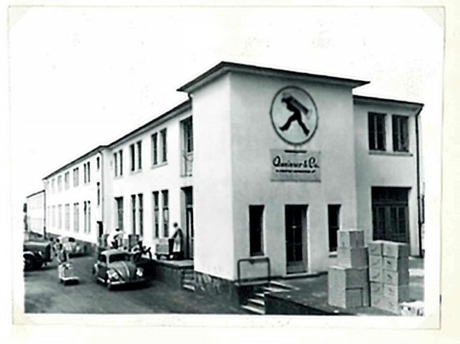En sort-hvid foto af en bygning med en stor logo af en person i bevægelse på facaden. Der står kasser foran bygningen, og der er nogle personer og biler i nærheden. Bygningens stil ser ud til at være fra midten af det 20. århundrede.