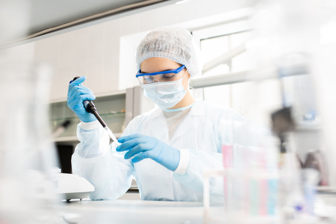 En laboratoriemedarbejder iført hvid kittel, ansigtsmaske og beskyttelsesbriller arbejder koncentreret med en pipette. Personen har blå handsker på og er omgivet af laboratorieudstyr.