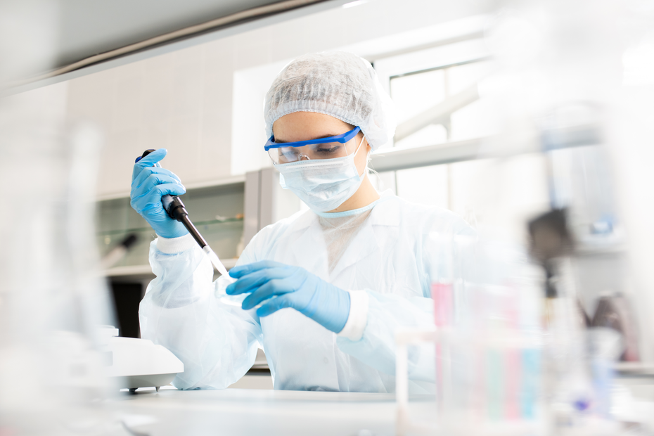 En laboratoriemedarbejder iført hvid kittel, ansigtsmaske og beskyttelsesbriller arbejder koncentreret med en pipette. Personen har blå handsker på og er omgivet af laboratorieudstyr.