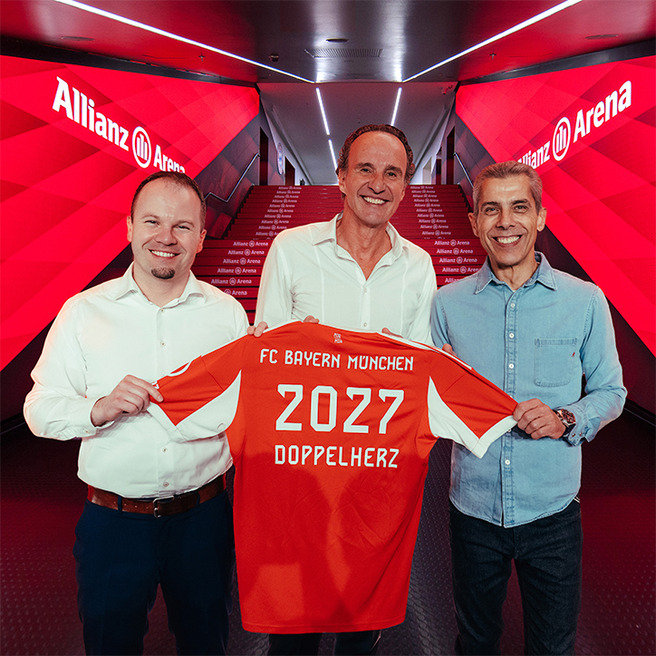 Tre personer står i en hal med lysrøde vægge. De holder en fodboldtrøje med teksten "FC Bayern München," årstallet "2027," og ordet "Doppelherz." Baggrunden viser en trappe med "Allianz Arena." Stemningen er festlig, og de smiler.