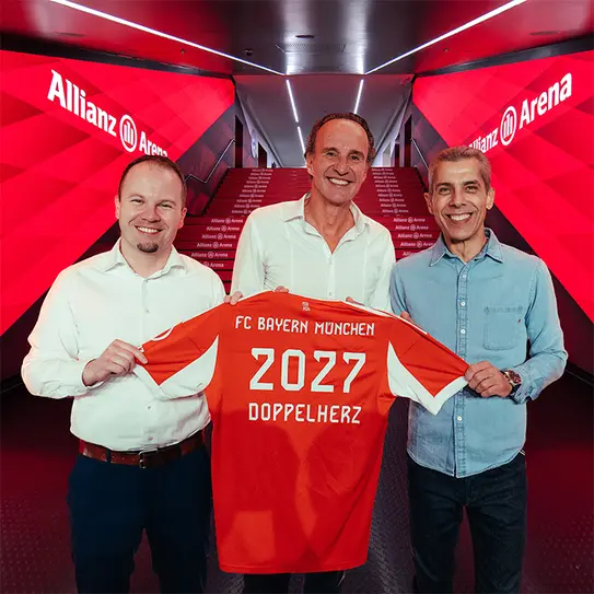 Tre personer står i en hal med lysrøde vægge. De holder en fodboldtrøje med teksten "FC Bayern München," årstallet "2027," og ordet "Doppelherz." Baggrunden viser en trappe med "Allianz Arena." Stemningen er festlig, og de smiler.