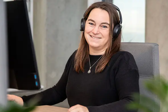 En smilende kvinde iført hovedtelefoner sidder ved et skrivebord med en computer. Hun har langt, brunt hår og bærer en sort trøje. I baggrunden ses en moderne kontorindretning.
