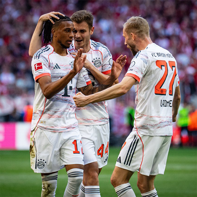 Tre spillere fra FC Bayern München fejrer en scoring. Den centrale spiller, iført trøje nummer 14, modtager lykønskninger fra sine holdkammerater, mens de udviser begejstring og sammenhold på banen. Stadionet er fyldt med fans i baggrunden, der bidrager til den intense atmosfære.