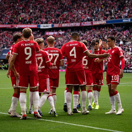 En gruppe fodboldspillere fra FC Bayern München i røde trøjer samles på banen, klapper hinanden og viser teamwork under en kamp. Publikum i baggrunden skaber en livlig atmosfære.
