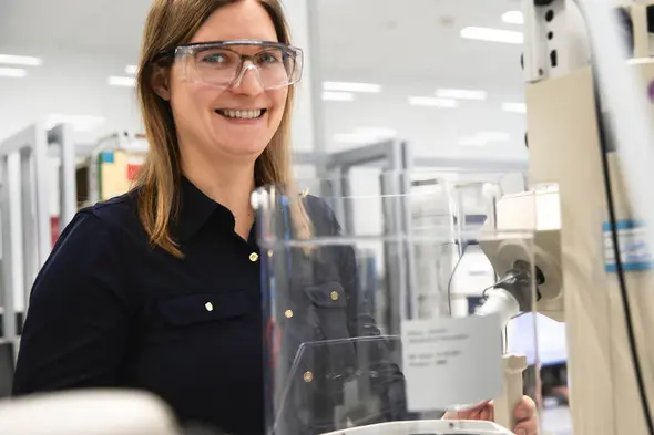 Kvinde i laboratorium bærer sikkerhedsbriller og smiler, mens hun står ved en laboratorieapparat. Baggrunden viser forskningsudstyr og en arbejdsstation.
