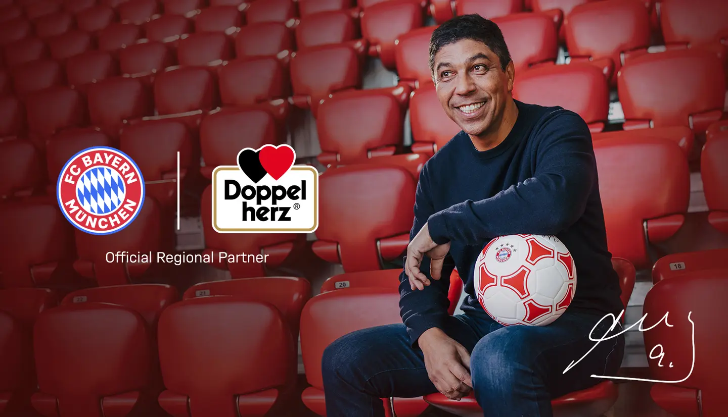 En smilende mand sidder på en række røde stadionpladser og holder en fodbold. Han er iført en sort sweater og ser venlig ud. I baggrunden ses logoerne for FC Bayern München og Doppelherz samt teksten "Official Regional Partner".
