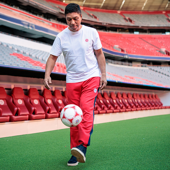 En mand står på en fodboldbane og spiller fodbold. Han har en hvid t-shirt og røde bukser. I baggrunden ses tilskuerpladserne i stadions. Manden koncentrerer sig om bolden, mens han dribbler den.