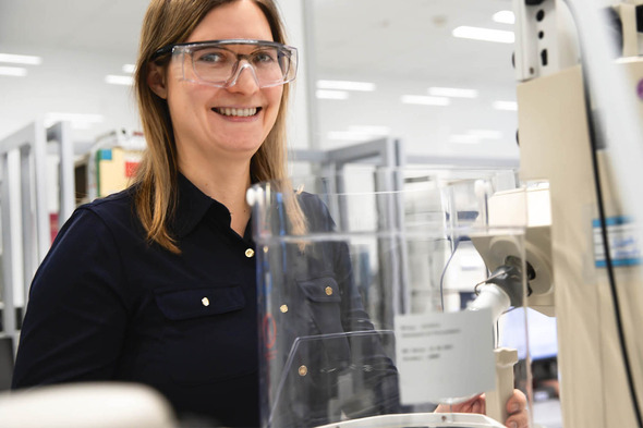 Kvinde i laboratorium bærer sikkerhedsbriller og smiler, mens hun står ved en laboratorieapparat. Baggrunden viser forskningsudstyr og en arbejdsstation.