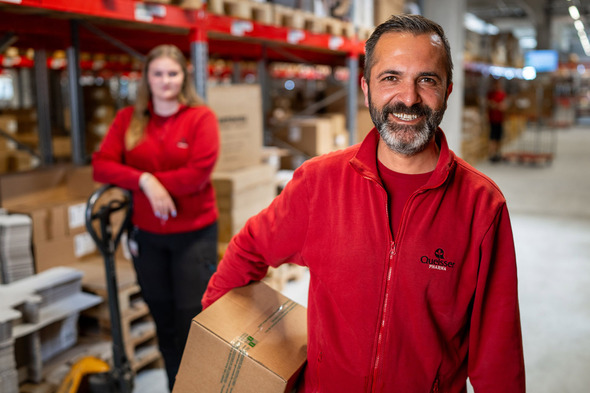 To mænd i røde arbejdsjakker står i et lagerområde. Den ene mand smiler, mens han holder en kasse, og den anden kvinde står i baggrunden med en palleløfter. Begge ser ud til at arbejde sammen i et travlt miljø.